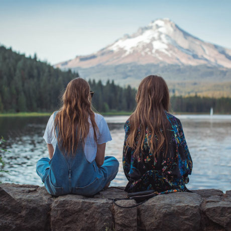 Two women sitting on a retaining wall next to a lake and mountains